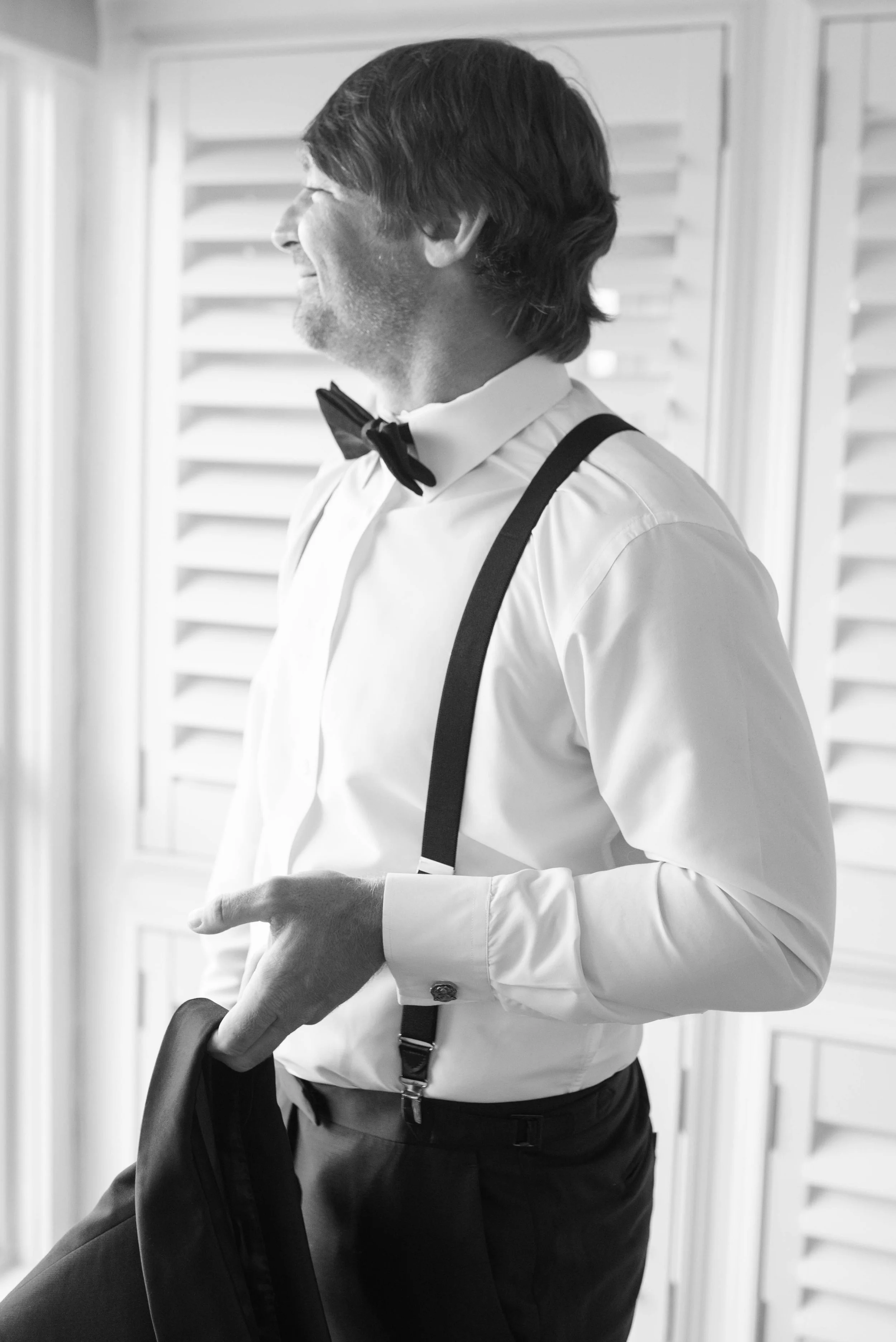 Side profile of the groom with medium-length dark hair, wearing a white dress shirt, black suspenders, and a black bow tie, holding a jacket in his hand, standing near wooden shutters in a room.