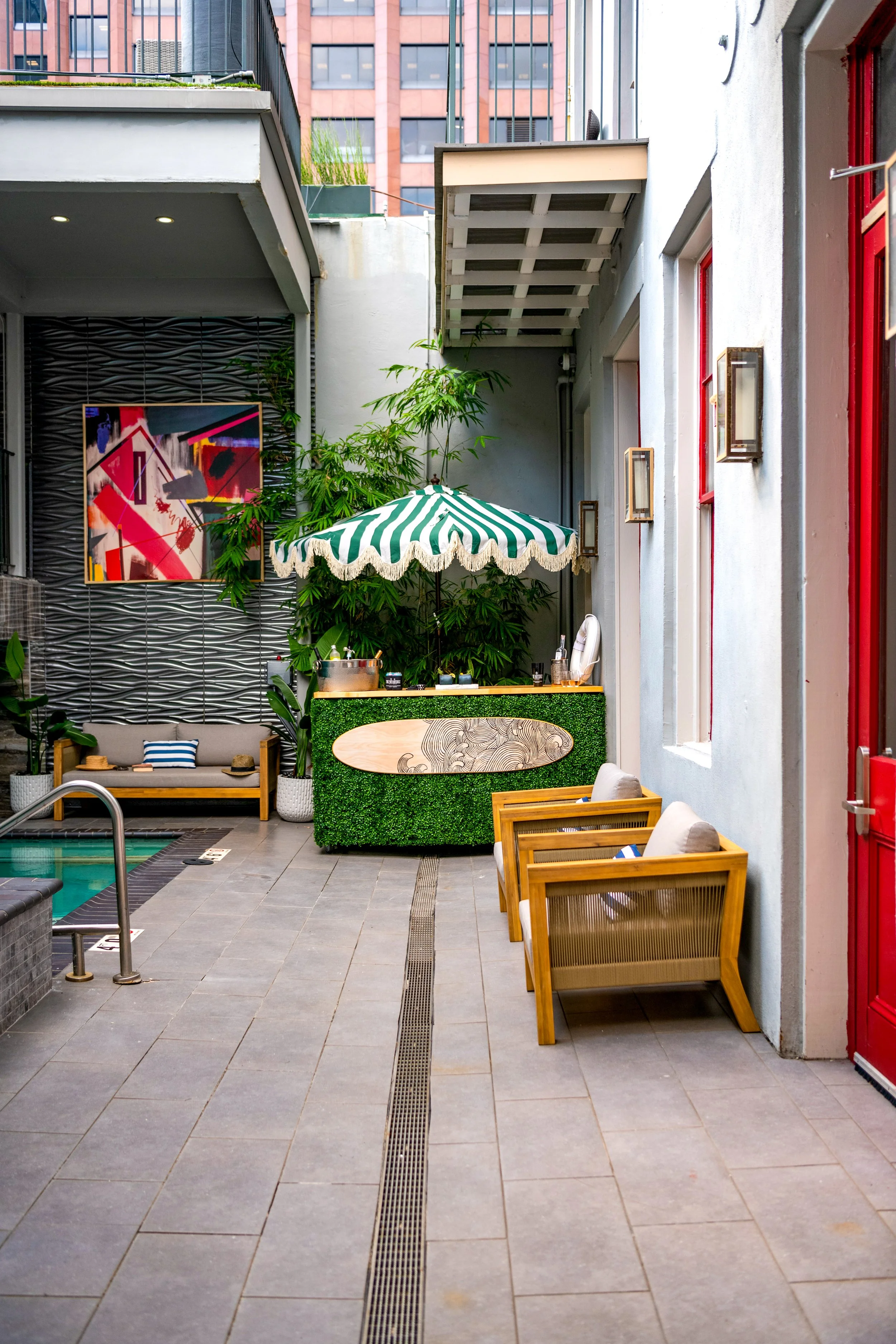 Outdoor seating area with wooden chairs with beige cushions along a gray tiled floor, a pool area on the left, a green decorated counter with a striped green and white umbrella, and a building with red windows and modern high-rise buildings in the background.