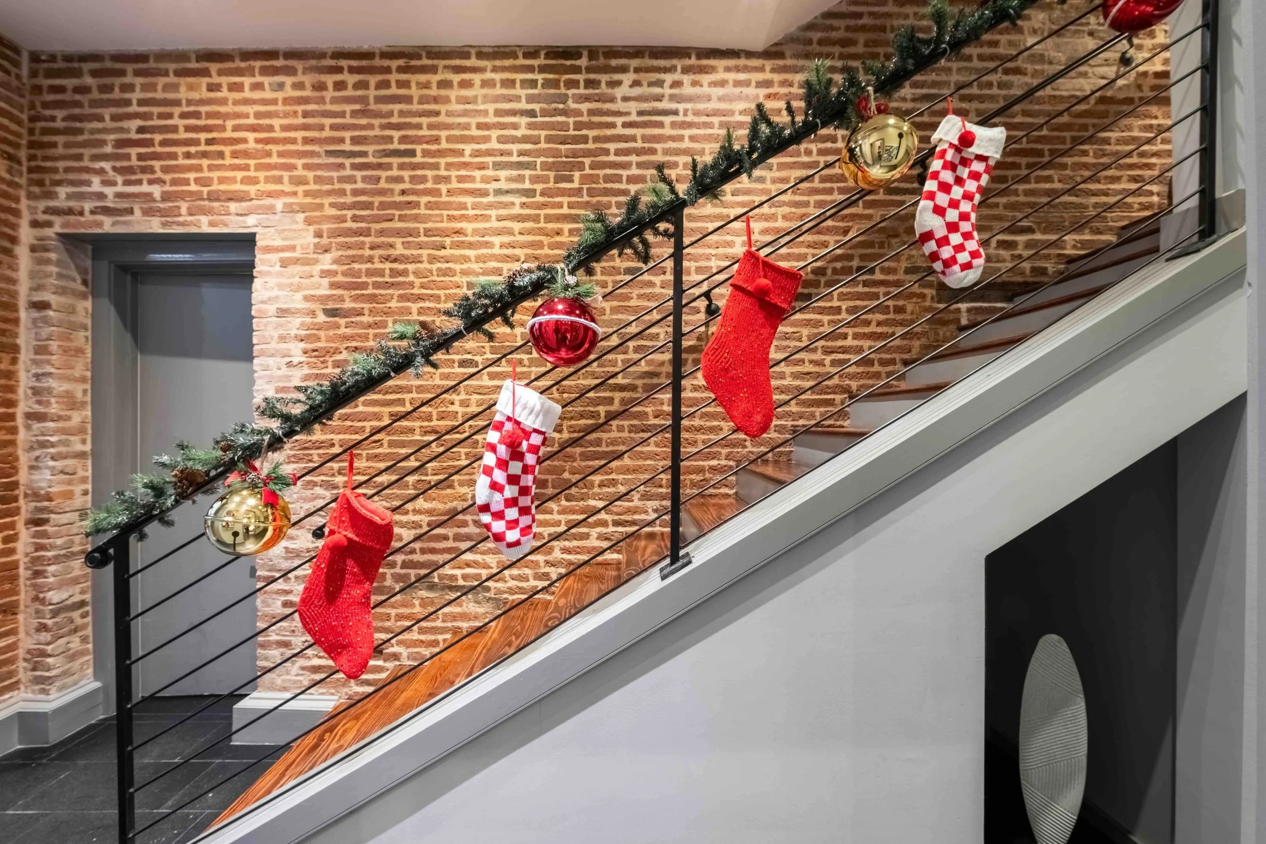 Christmas stockings and ornaments hanging on a decorated railing along a staircase with brick wall background.