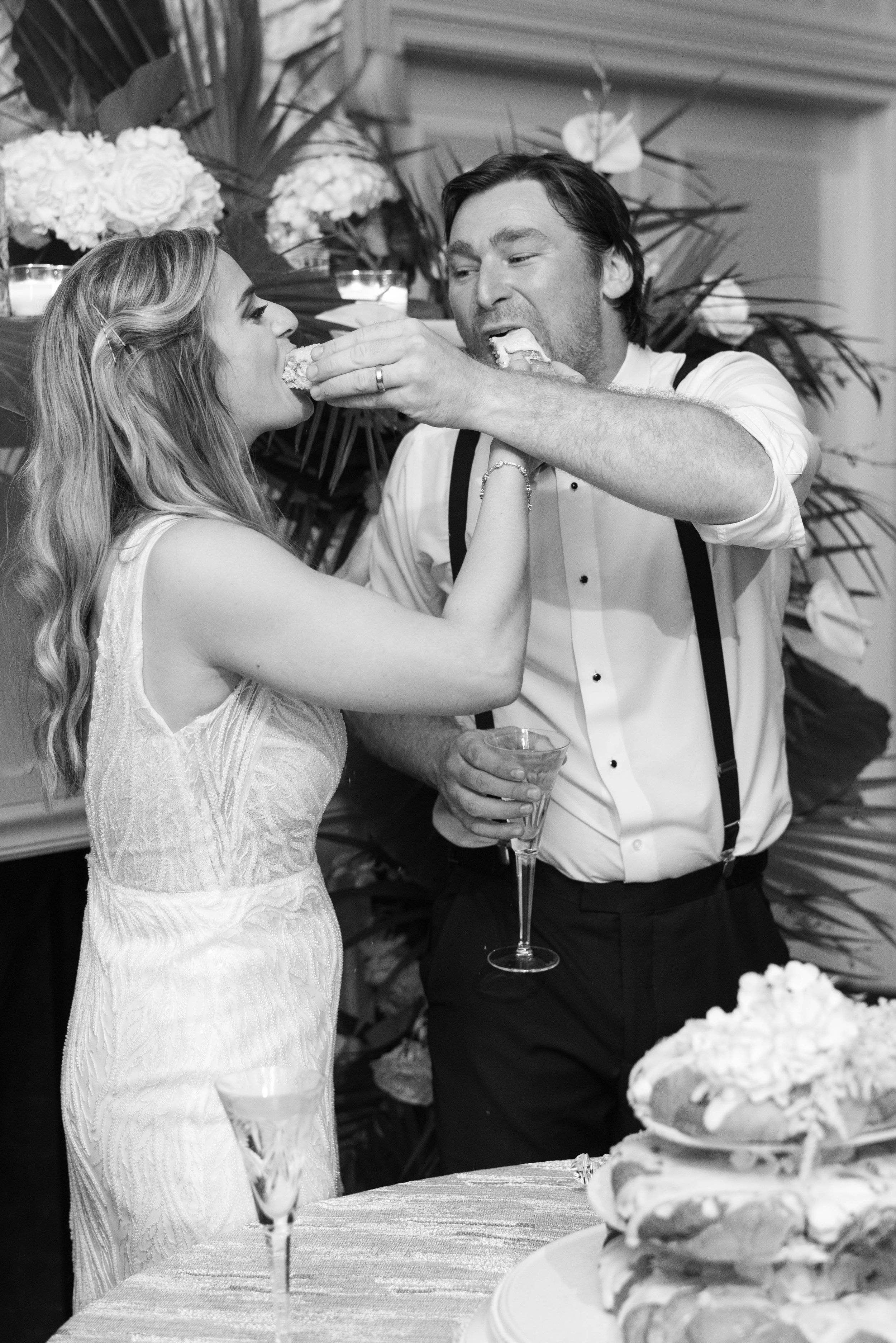 A black and white photograph of a man feeding a woman a piece of cake at a wedding, with a large layered cake in the foreground and floral decorations in the background.
