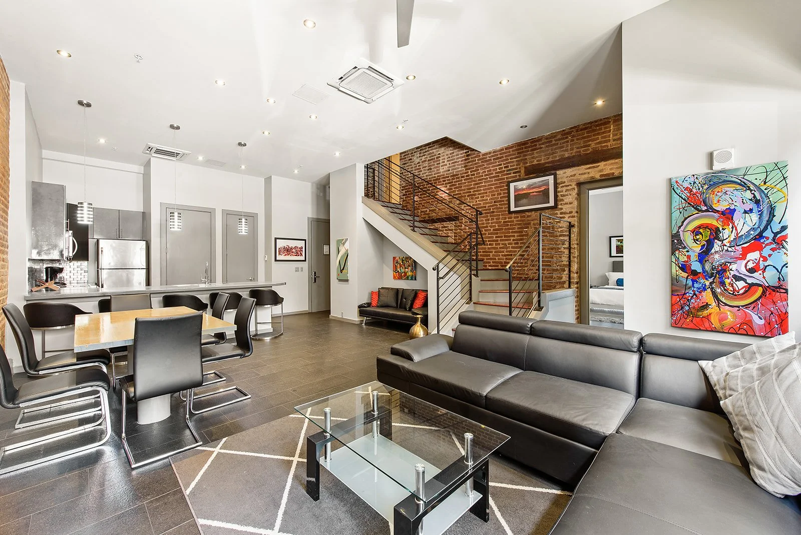Modern open-concept living room and kitchen with brick accent wall, black leather sofa, abstract artwork, dining table with black chairs, and staircase.