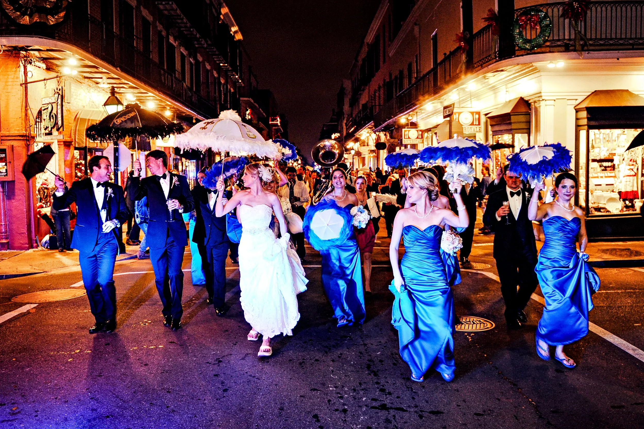 A nighttime street scene with a group of people in formal attire, including wedding dresses and tuxedos, walking down the street in a wedding 2nd line with umbrellas and smiling, surrounded by shops and streetlights.