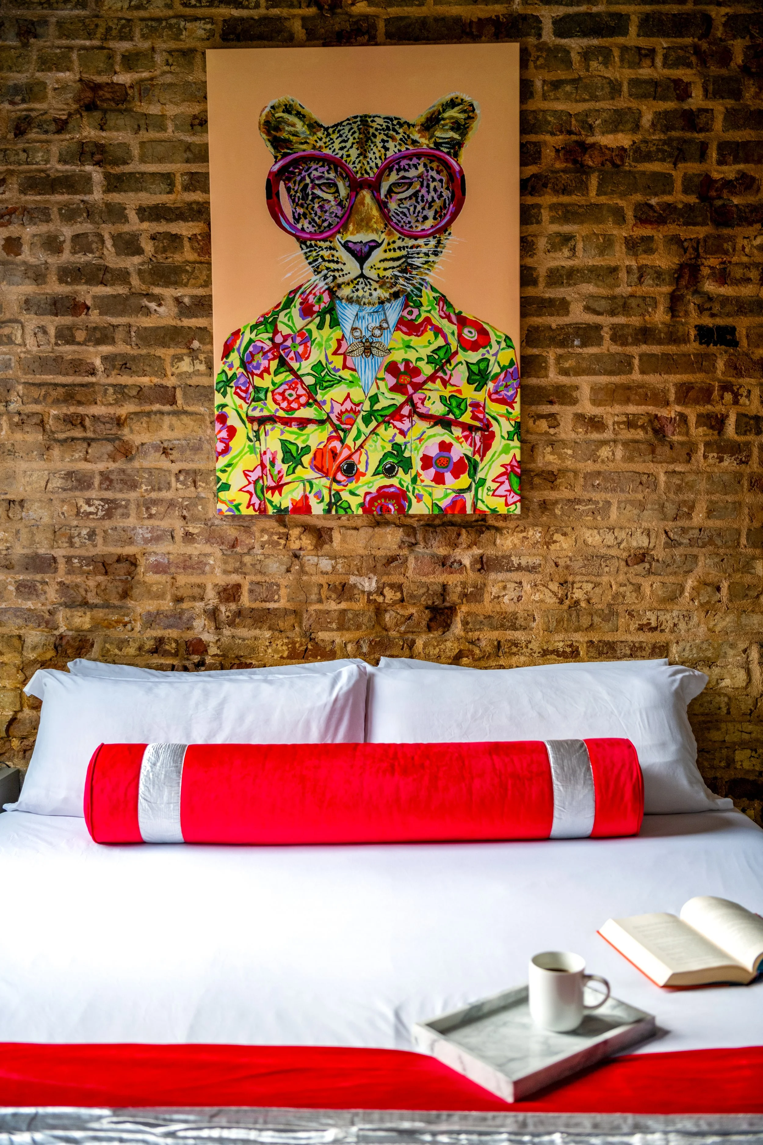 A bed with white sheets and pillows, decorated with a red and white cylindrical bolster pillow, in front of a brick wall. Above the bed hangs a colorful painting of a leopard wearing pink glasses and a floral jacket. On the bedside table, there is a tray holding a white mug, an open book, and a small cup.