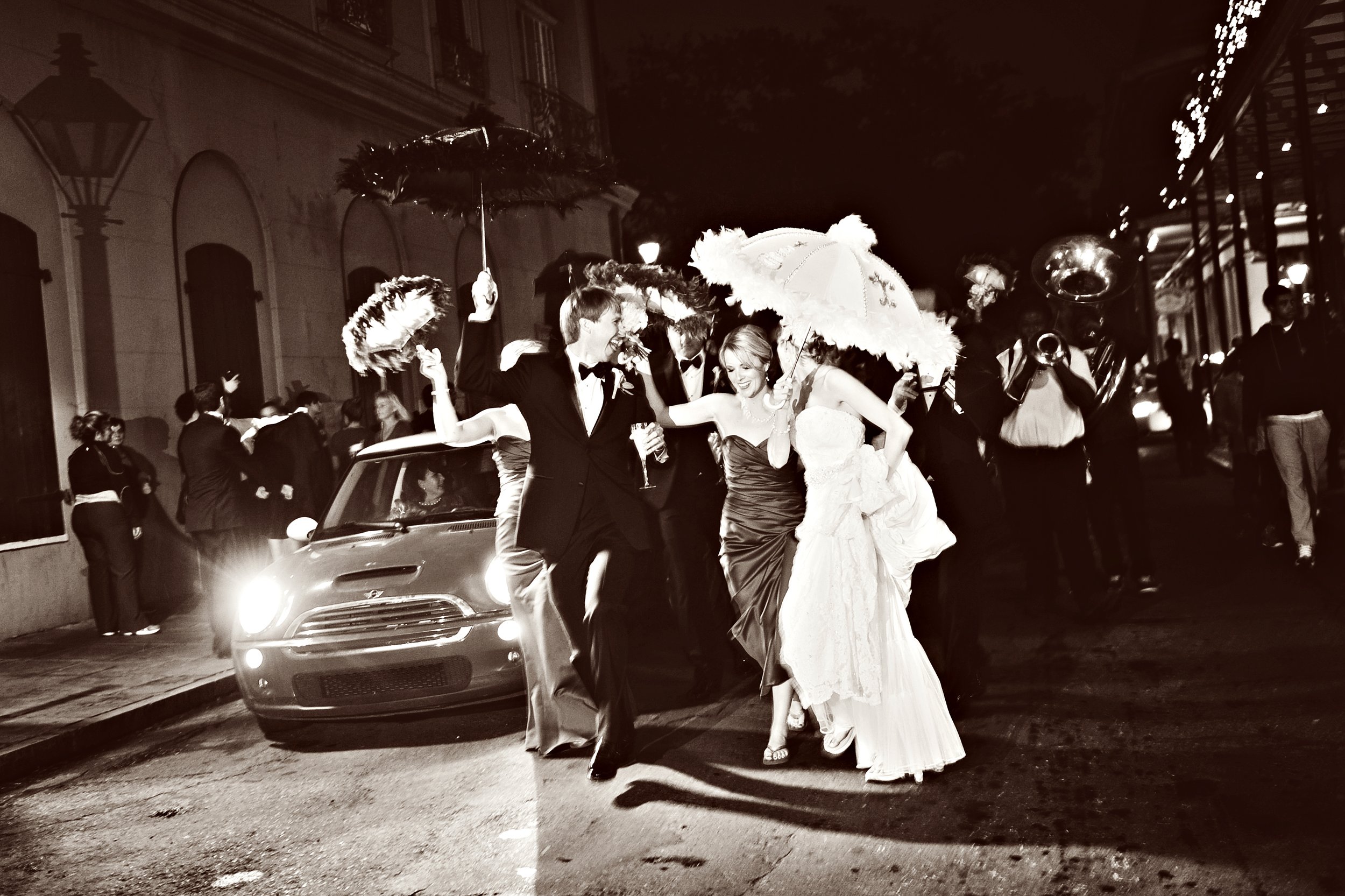 Nighttime scene of a wedding celebration with a bride and groom walking together in a second line procession, surrounded by a group of people, some playing musical instruments, and others holding umbrellas.