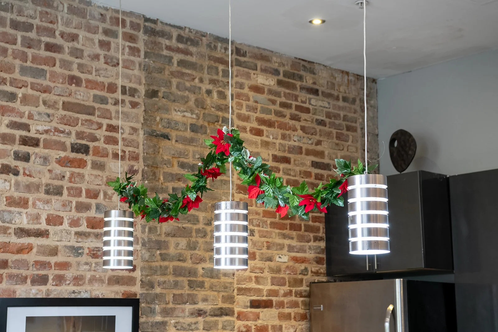 Three hanging pendant lights decorated with a Christmas garland with artificial holly leaves and red poinsettia flowers, in a modern kitchen with a brick wall.