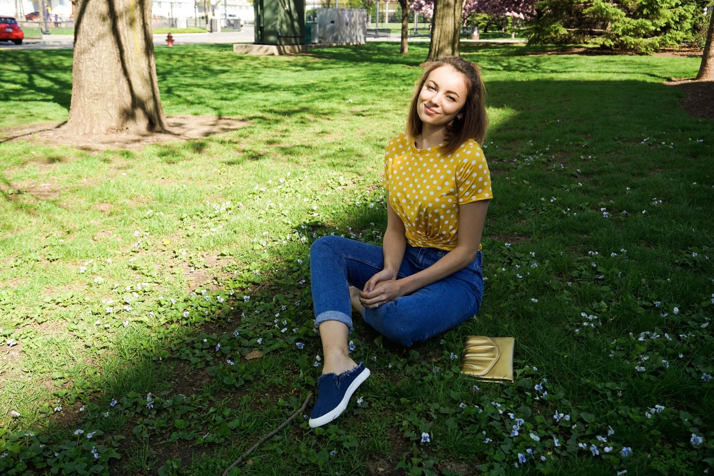 Blogger sitting on the grass, wearing a yellow and white polka dot crop top, mom jeans, denim slip ons, and a golden clutch.