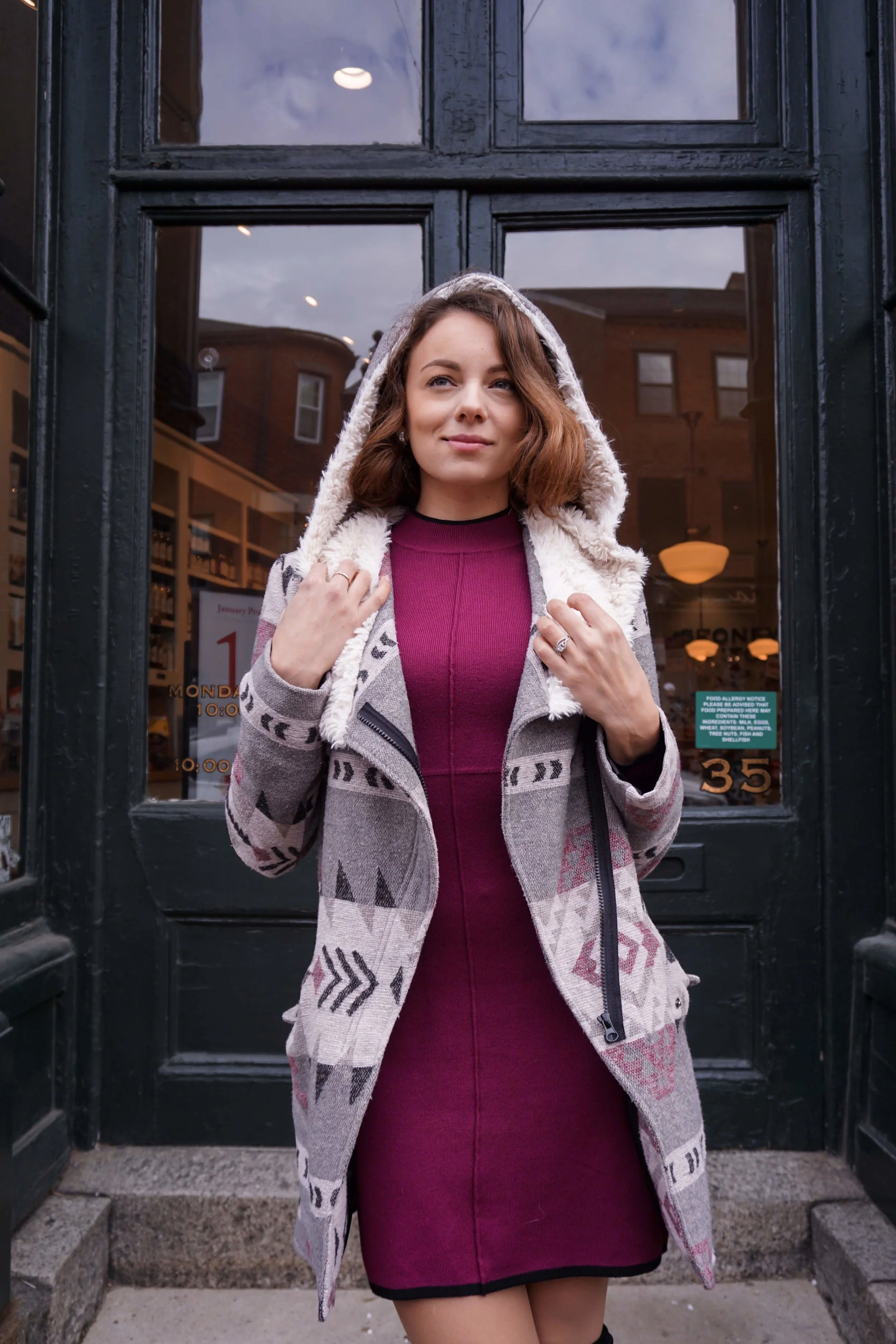A blogger posing on the street wearing a maroon dress, cozy coat, knee-high boots.