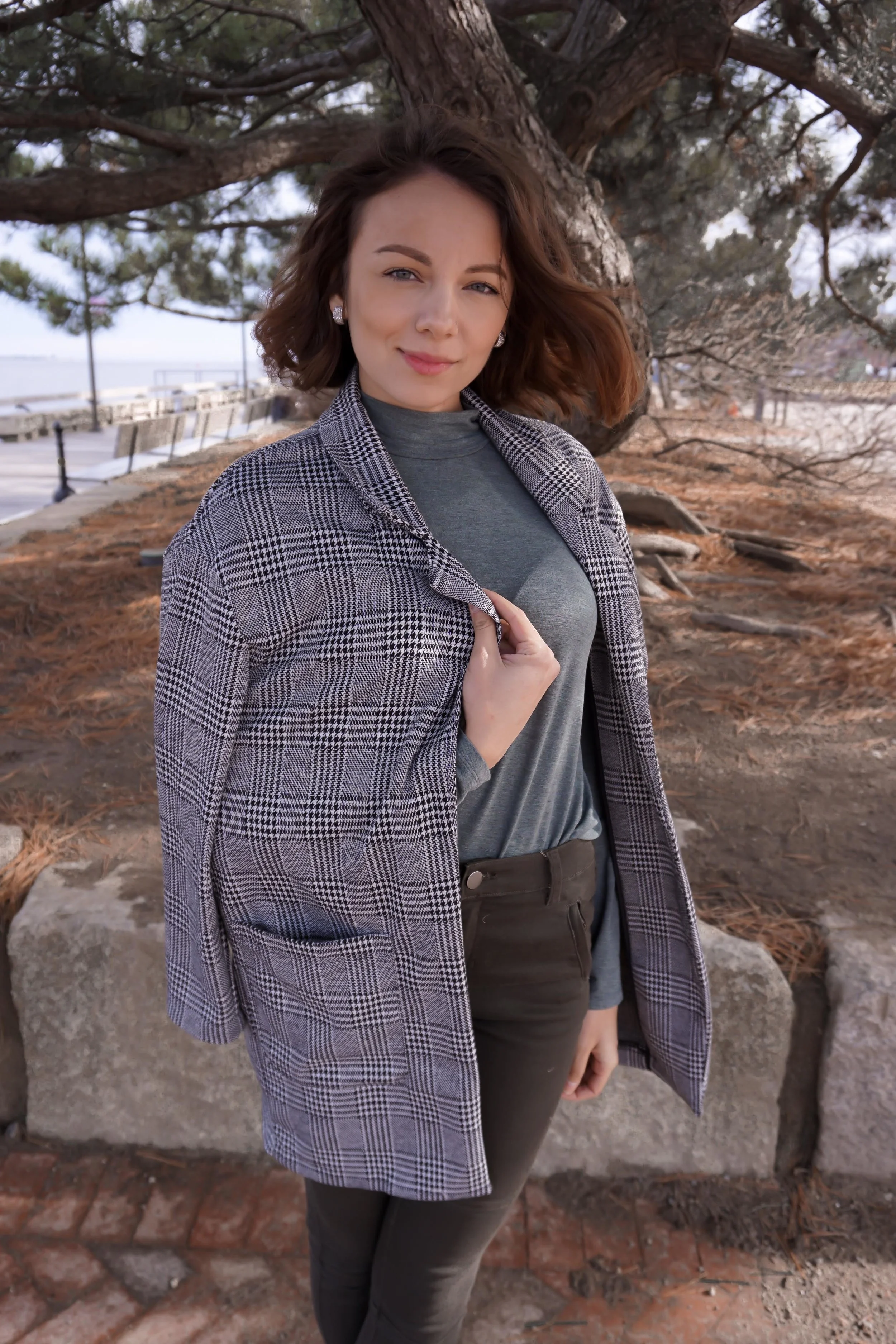 A blogger posing near the water, wearing camel green ankle pants, turtleneck, black point heels, and a houndtooth blazer.
