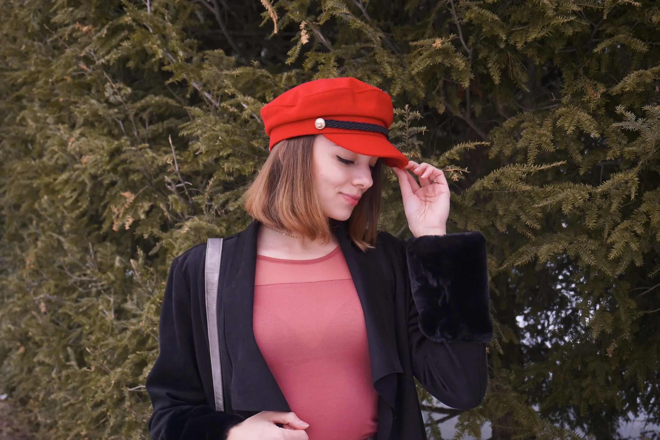 Fashion blogger is posing in the nature, wearing a red hat, salmon top, black jacket, gray pencil skirt, and faux-leather boots.