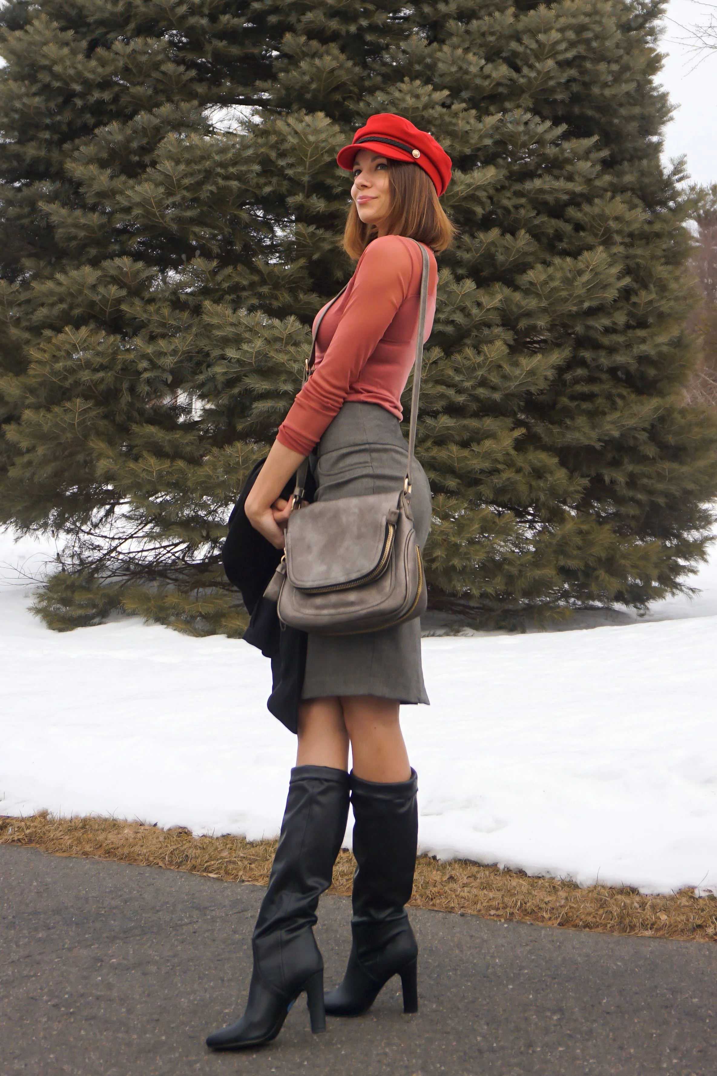 Fashion blogger is posing in the nature, wearing a red hat, salmon top, black jacket, gray pencil skirt, and faux-leather boots.