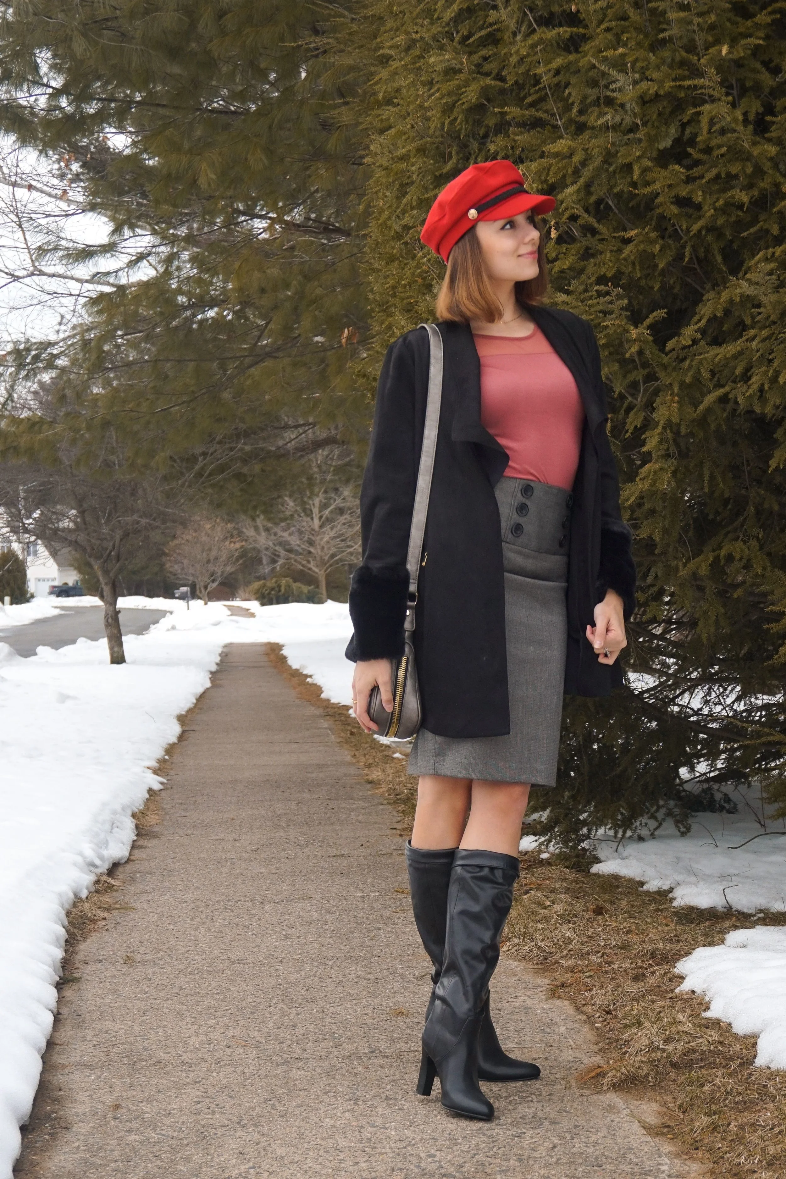 Fashion blogger is posing in the nature, wearing a red hat, salmon top, black jacket, gray pencil skirt, and faux-leather boots.