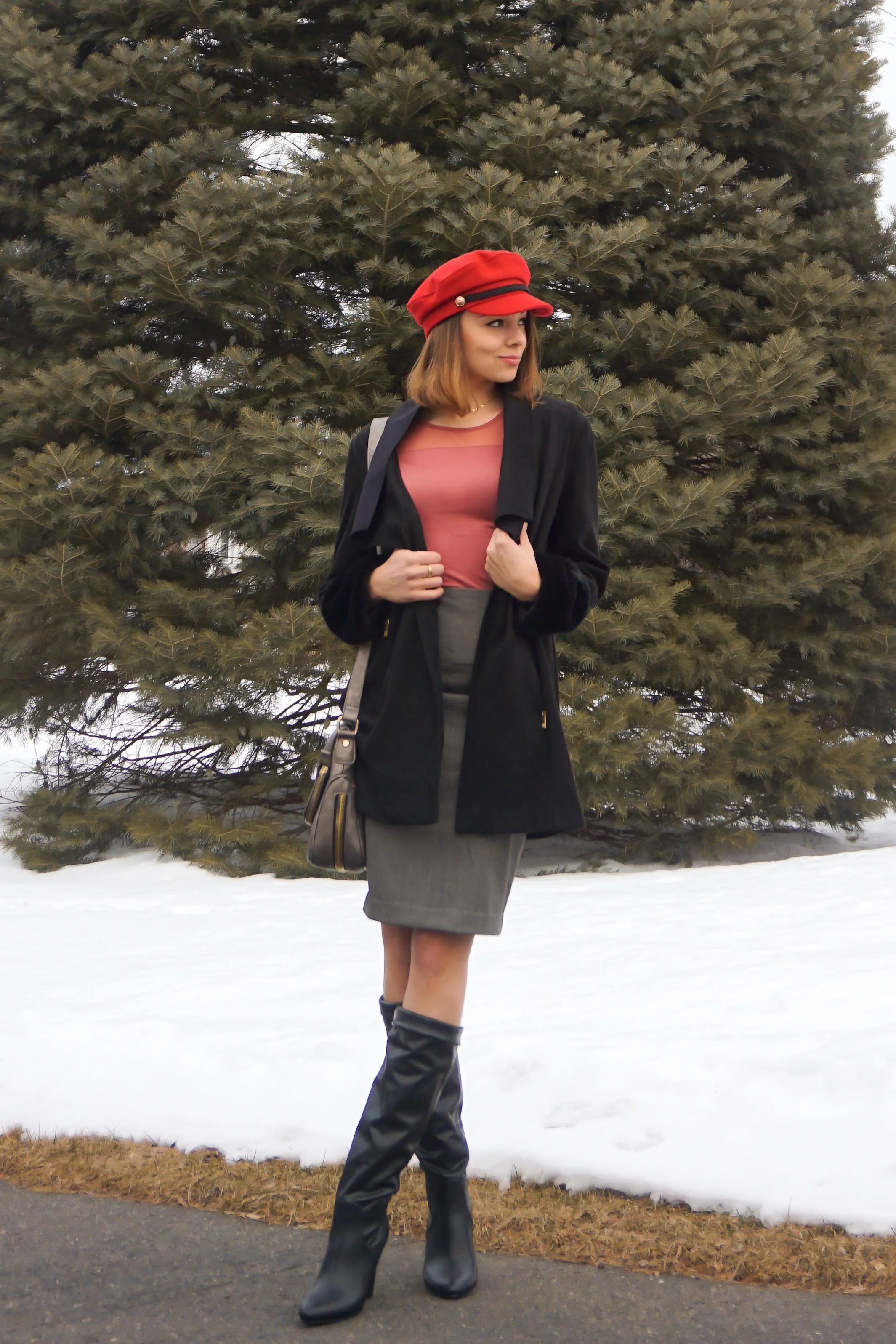 Fashion blogger is posing in the nature, wearing a red hat, salmon top, black jacket, gray pencil skirt, and faux-leather boots.