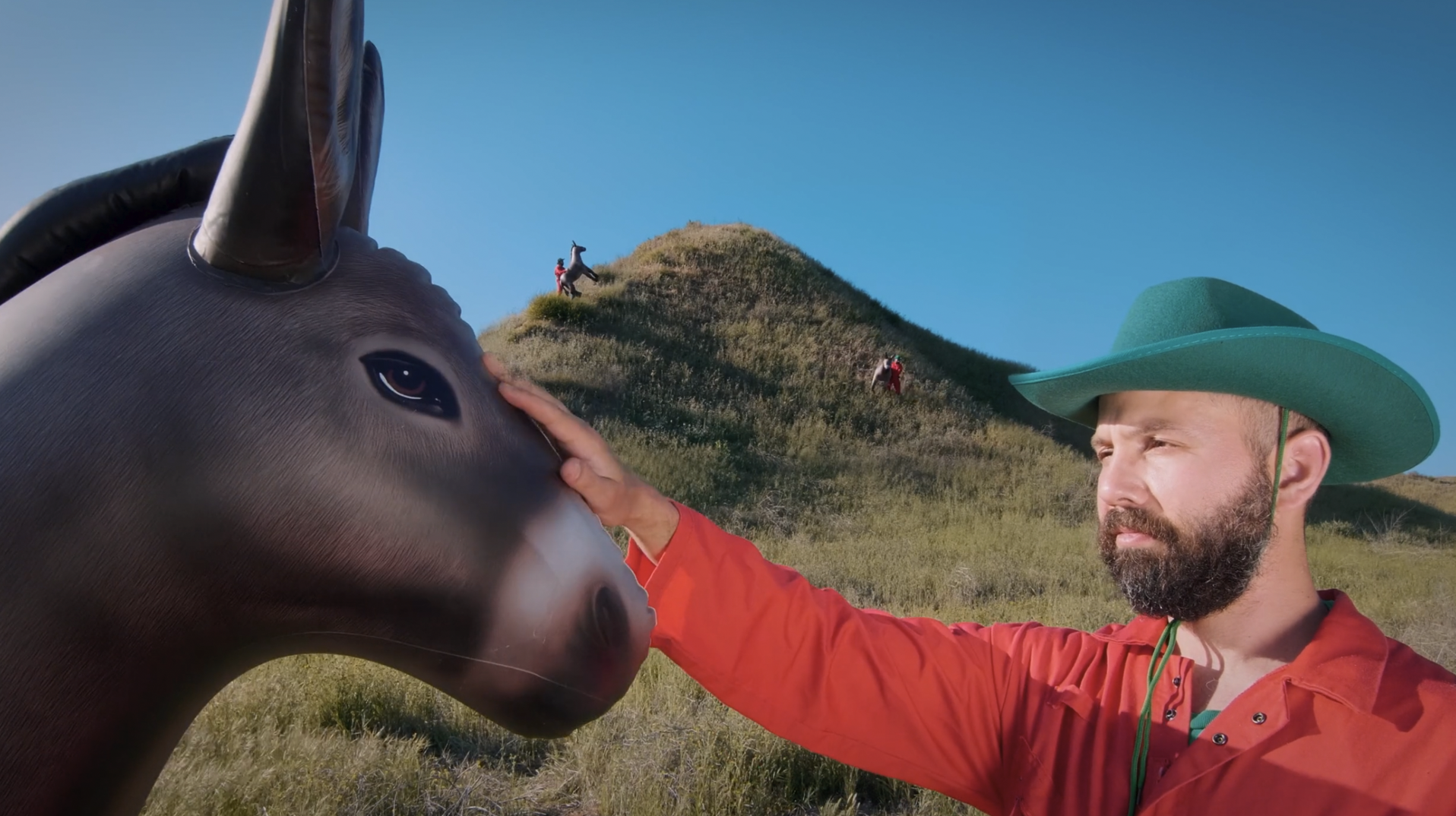 Jack Fleischer as J4CKSN4CK pats the head of an inflatable donkey in front of a rolling green hill and blue skies