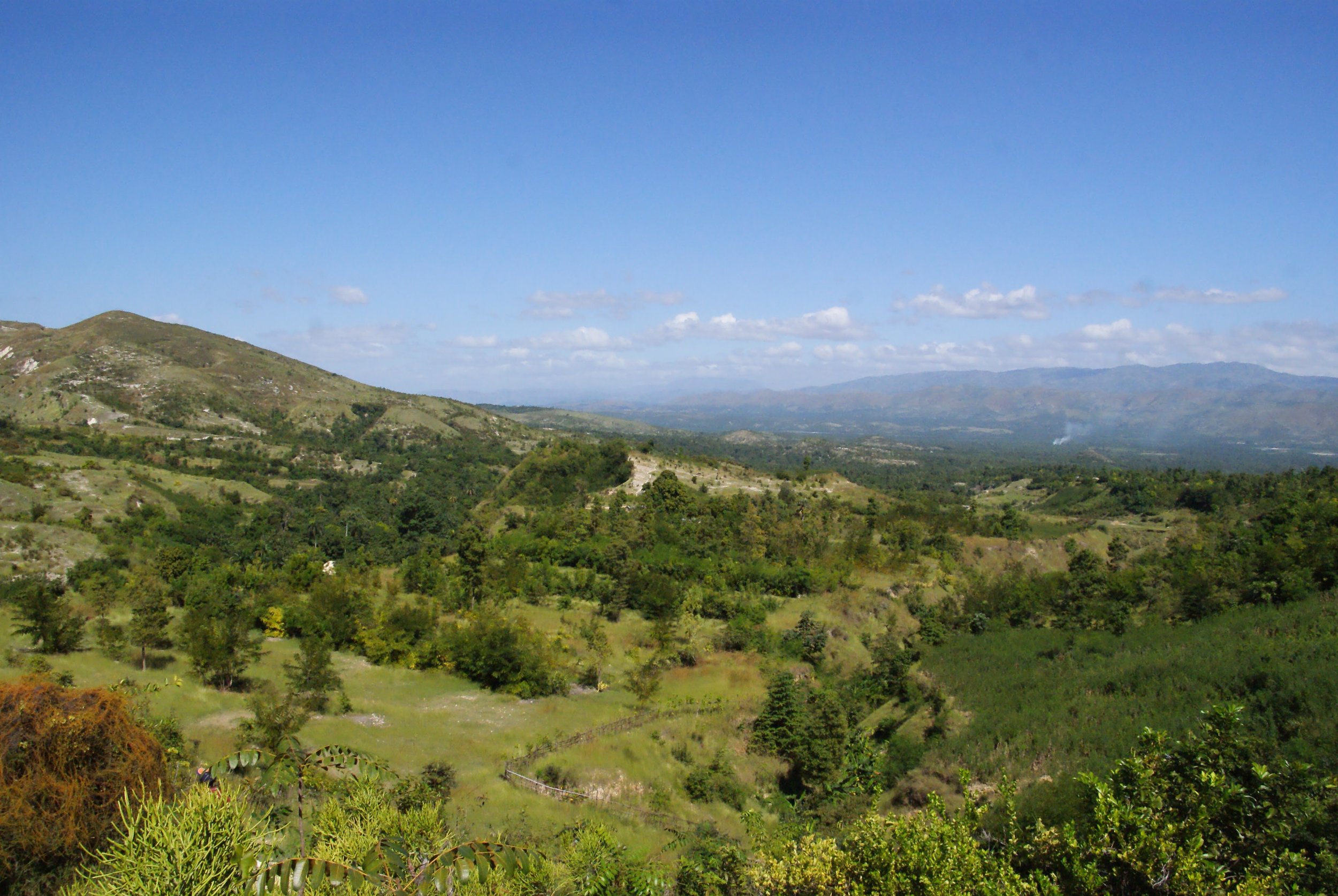 MCC has supported the reforestation work in the Artibonite Valley that is visible in the middle of this photo. Prior to reforestation, agro-forestry and micro-forestry work, the mountainside was as bare as the mountain in the upper left of the photo. Photo credit: Tina Schrag