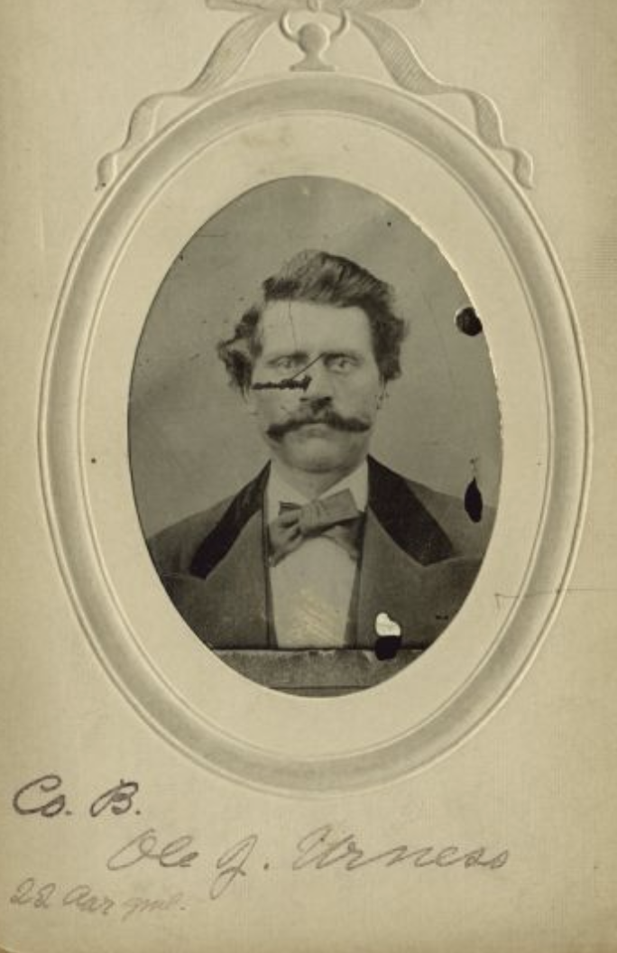 Black and white photograph of a man with a mustache and sideburns, wearing a suit with a bow tie, framed in an oval with decorative border, with handwritten text below.