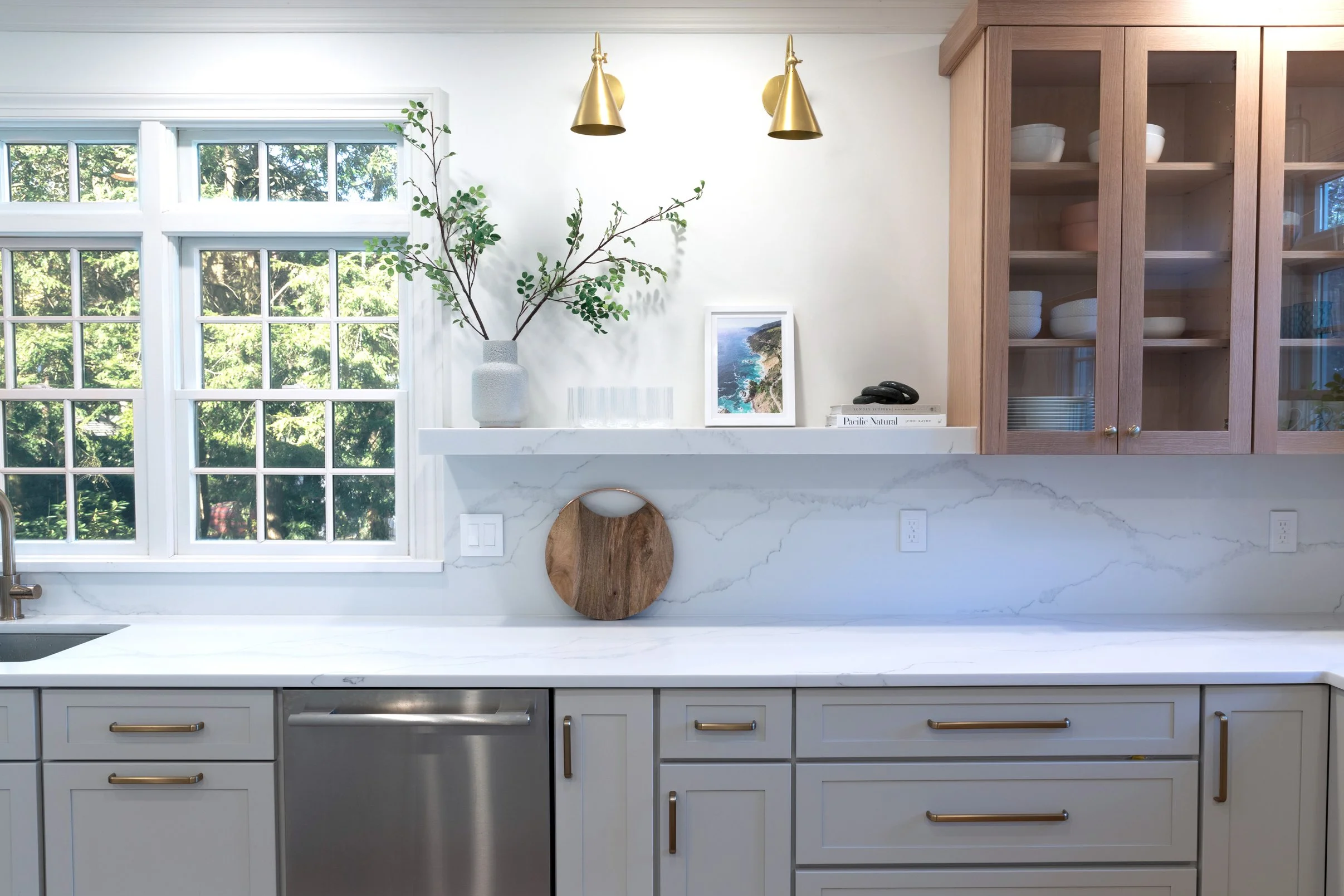 Bright kitchen with white cabinets and marble countertops, wooden glass-front cabinets, a large window showing green trees, and decorative items including a vase with greenery, framed photo, glassware, and books.