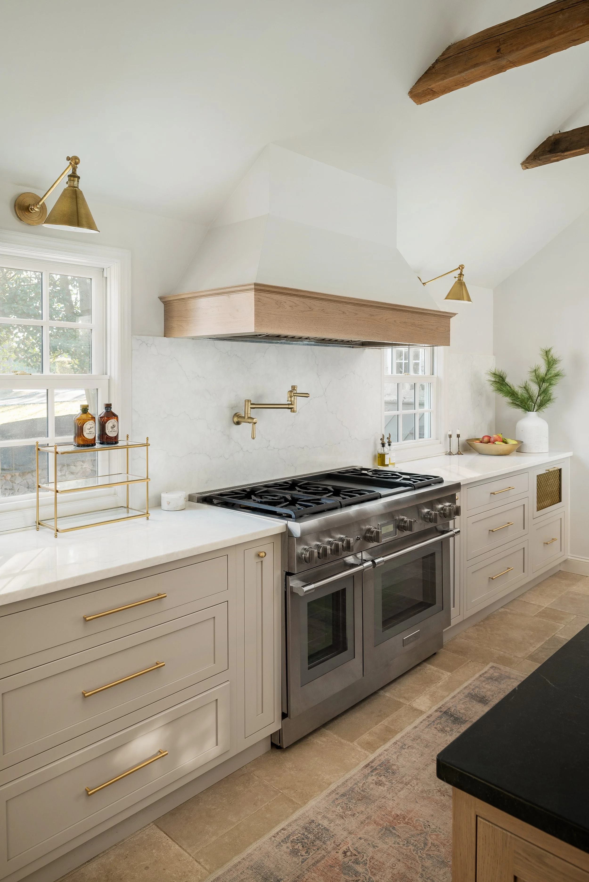 Kitchen with white cabinets, marble backsplash, stainless steel double oven, black countertop, window, gold fixtures, wooden beams on ceiling, and decorative items.