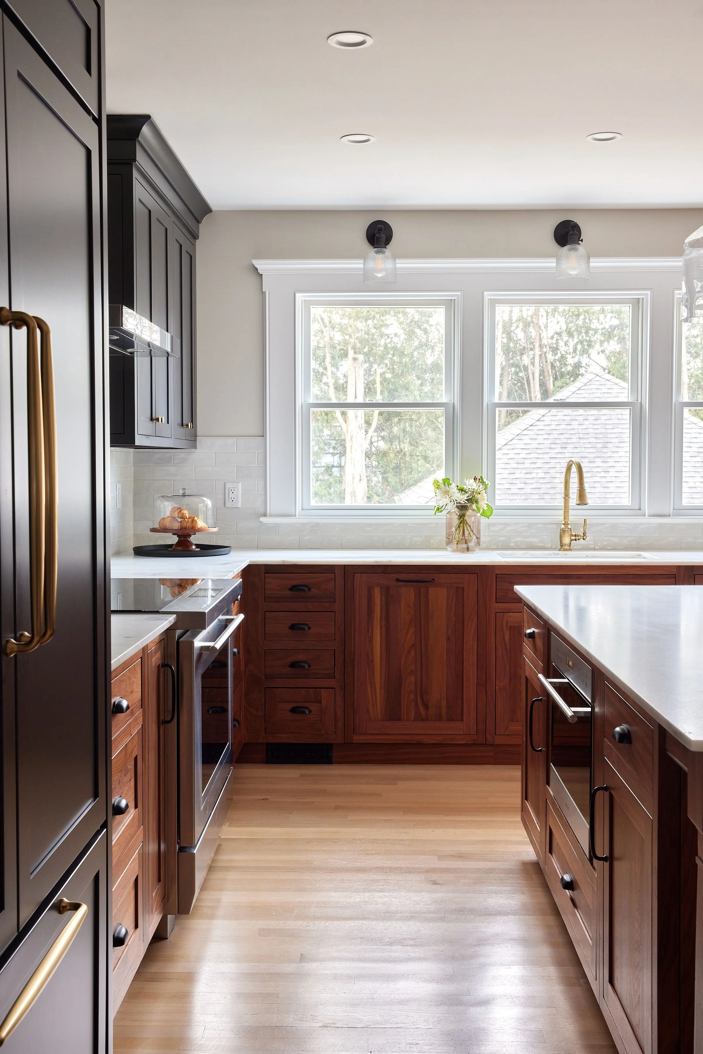 A modern kitchen with wooden cabinets, a white countertop, large windows, a gold faucet, and minimal decorative elements.