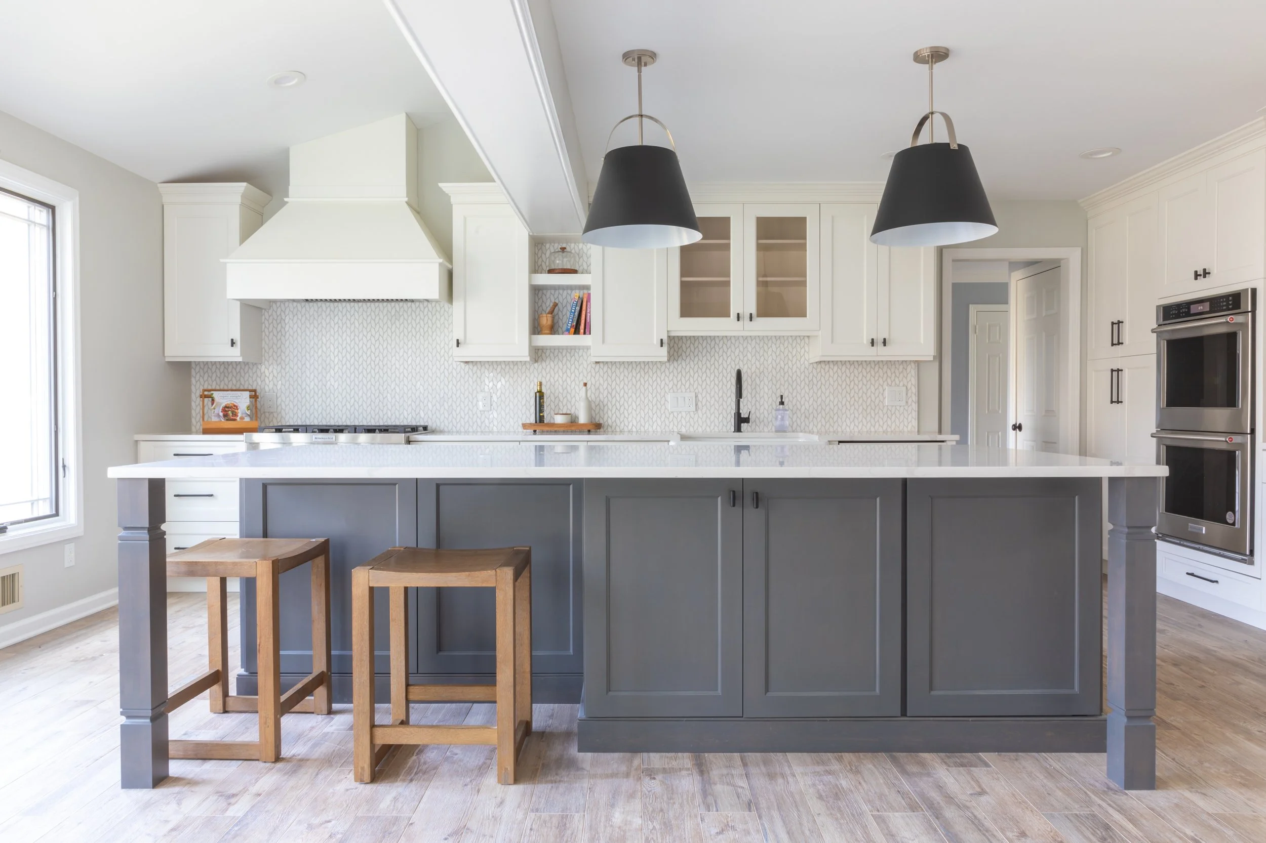 Modern kitchen with white upper cabinets, dark grey lower cabinets and island, two wooden bar stools, pendant lights, and stainless steel double oven.