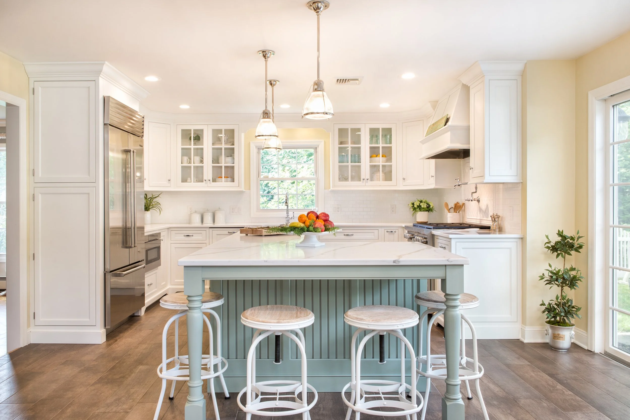 Bright white kitchen with central island, four chairs, and a bowl of fruit, featuring white cabinets, windows, and hardwood floors.