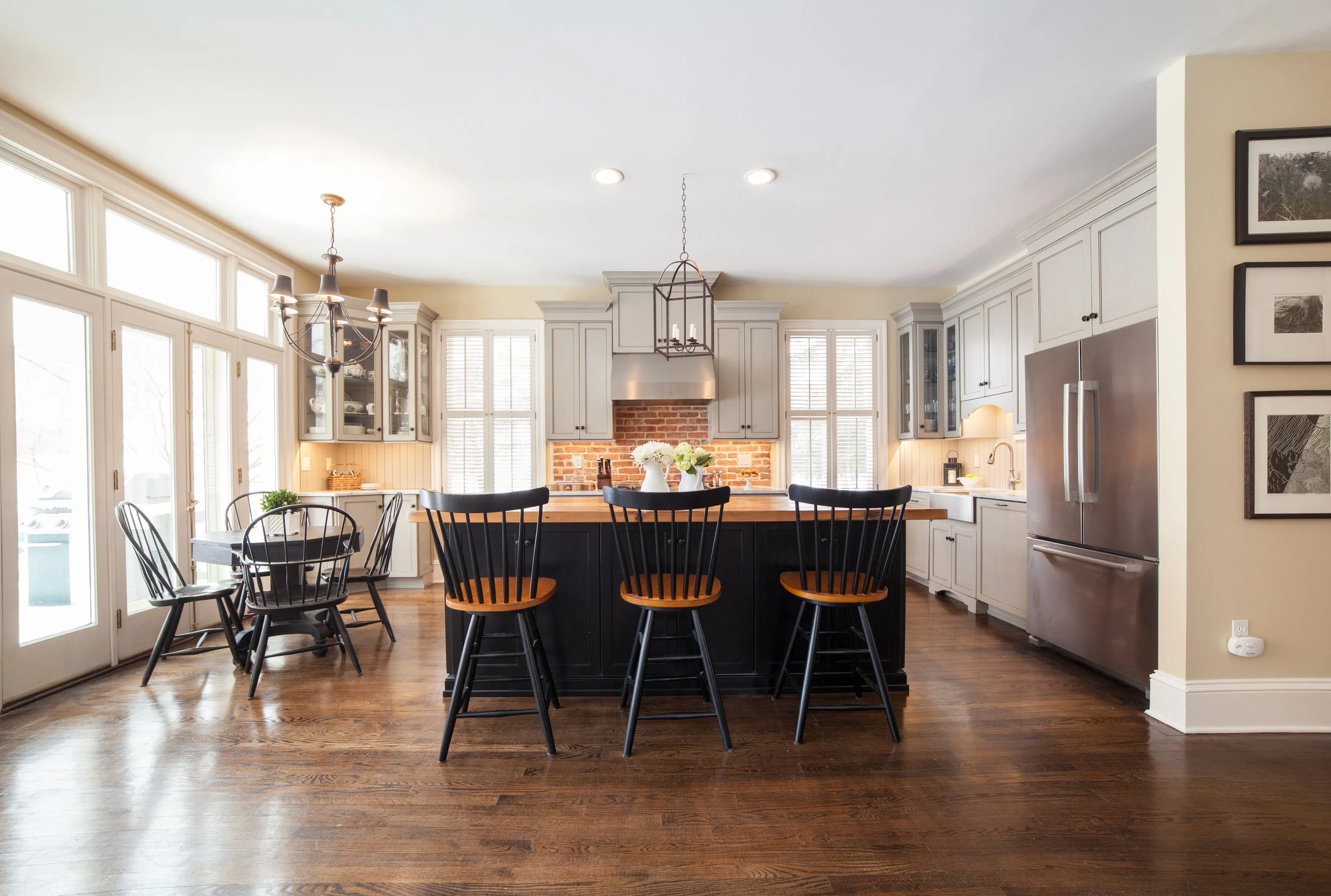 Bright kitchen with white cabinetry, a black island with wooden barstools, a dining table with chairs, stainless steel refrigerator, brick backsplash, and large windows.