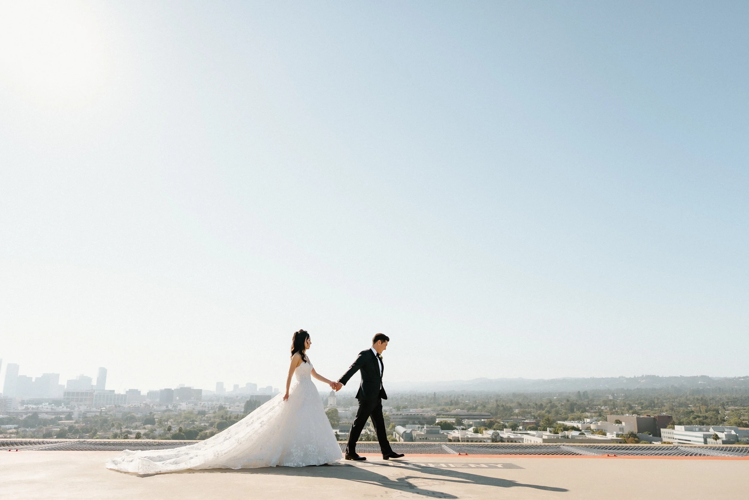 Bride in Pronovias ballgown cascading behind her and Groom walk across Helipad on Four Seasons Hotel Los Angeles at Beverly Hills with Los Angeles Skyline in the background