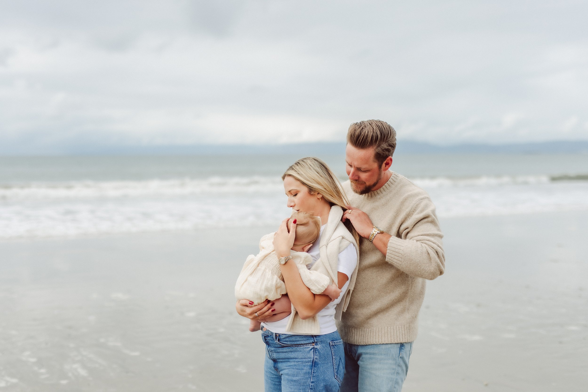 Husband embraces wife holding infant on the beach in Palos Verdes California