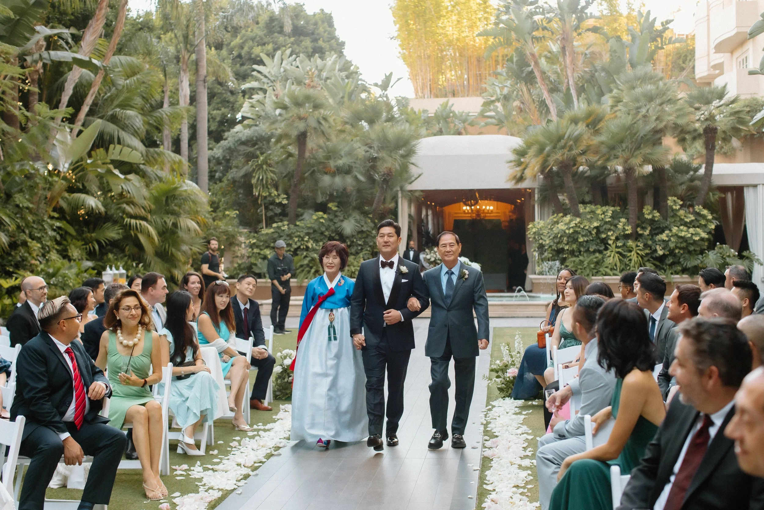 Groom and his mother in traditional Korean Hanbok and father in blue suit during wedding processional in the garden at Four Seasons Hotel