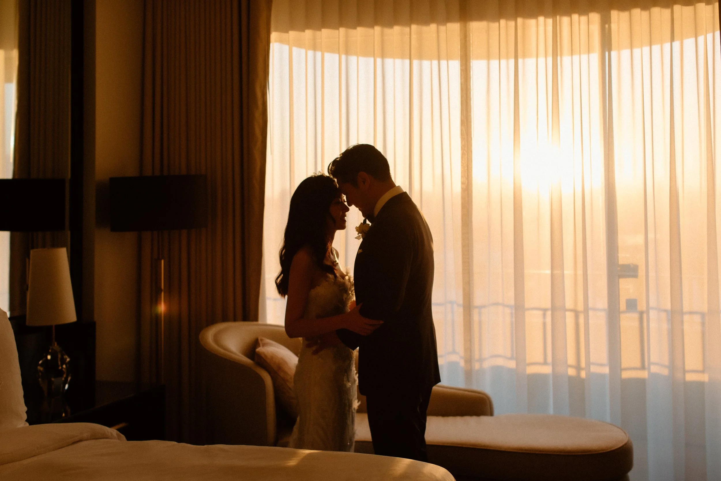 Bride and groom embrace in their wedding suite of Four Seasons Hotel with sunset flooding through the floor to ceiling windows. 