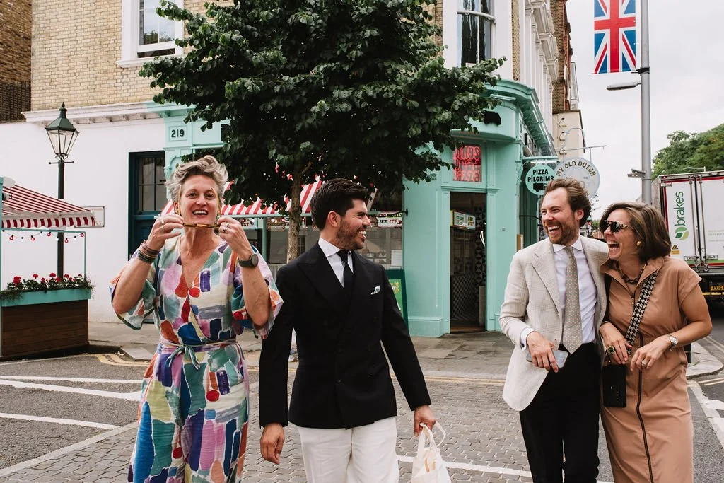 Four people with light skin walking on a city street, laughing and smiling. One woman is eating a hot dog. The background has a building with a mint green storefront, a UK flag, and outdoor decor.