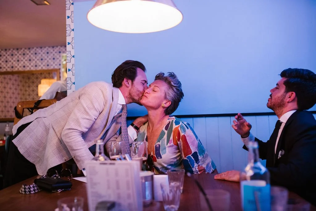 A man in a beige suit kisses an older woman on the cheek at a dinner table, with two men in tuxedos sitting nearby. The scene takes place in a restaurant with warm lighting and patterned walls.