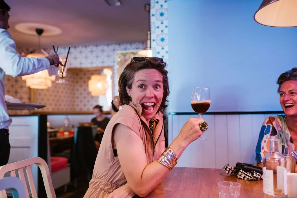 A woman with short brown hair, wearing a beige sleeveless top, holding a glass of red wine, smiling excitedly at the camera in a lively restaurant or bar setting.