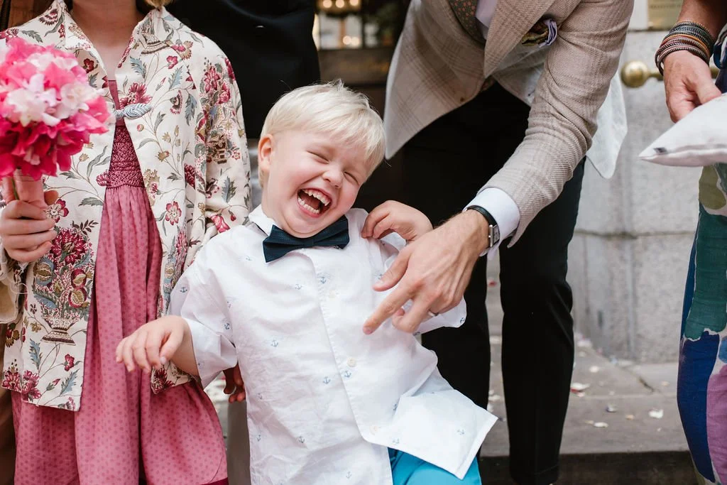 A young boy dressed in a white shirt and a bow tie is laughing joyfully while being touched on the shoulder by an adult man. To his left, a girl in a floral jacket holds a bouquet of pink flowers. The scene seems to be part of a celebration, possibly a wedding or special event, outdoors on a paved area.