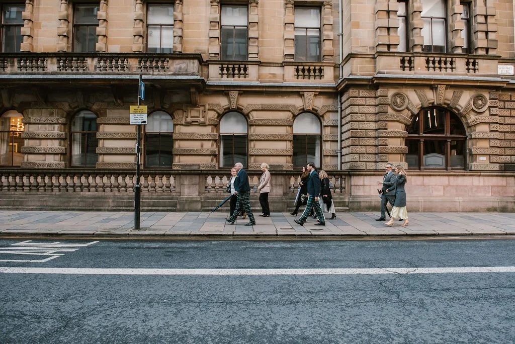 People walking on a city sidewalk in front of a historic stone building with arched windows.