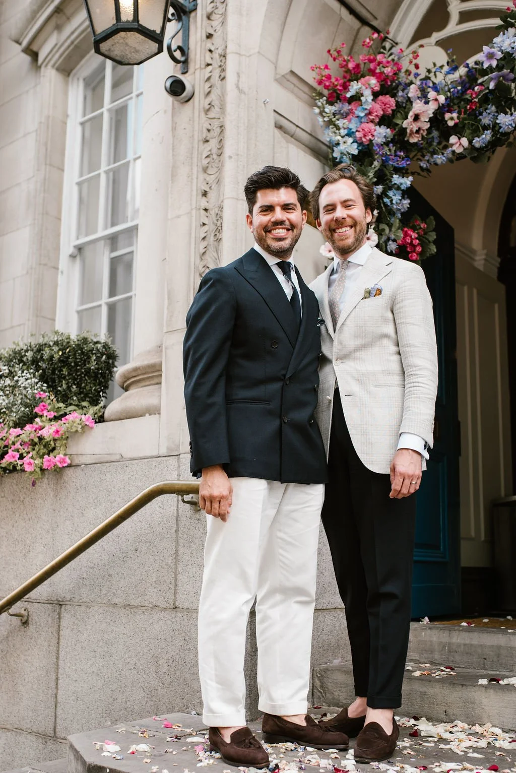 Two men in suits standing outside a historic building decorated with flowers, smiling at the camera.