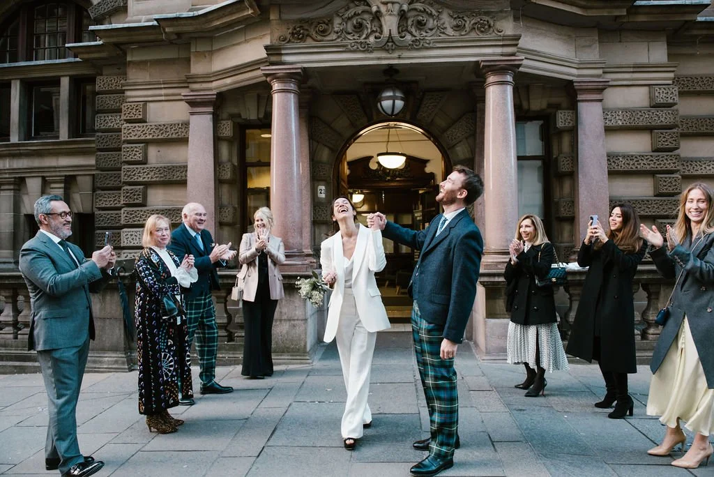 Bride and groom holding hands, smiling, walking out of a building, surrounded by guests clapping and taking photos.