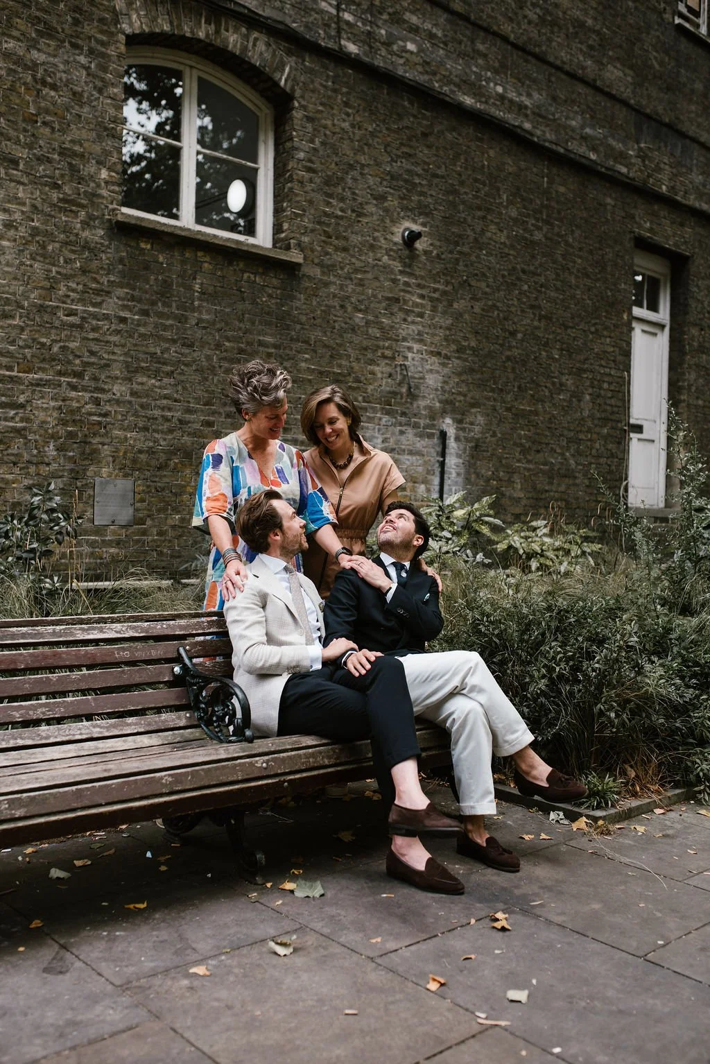 Two men sitting on a park bench, one in a beige suit and the other in a dark suit, are looking at each other and smiling. Two women, standing behind them, are smiling and looking at the men. The scene is set outdoors against a brick building wall with windows and greenery.