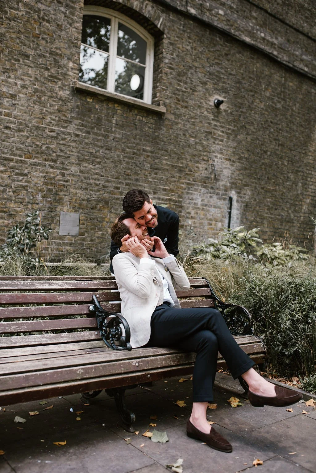 A woman and a man sharing a joyful moment on a park bench, with the woman sitting and the man standing behind her, holding her face and smiling.