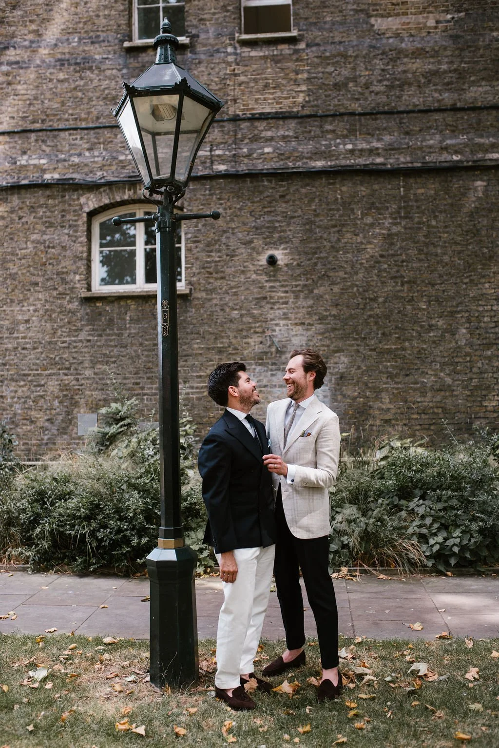 Two men in formal attire standing close and smiling at each other near a vintage street lamp, with a brick building and greenery in the background.