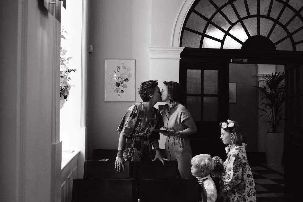 Two women kiss at a dining table while two young girls stand nearby in a room with large arched window, potted plant, and wall art.