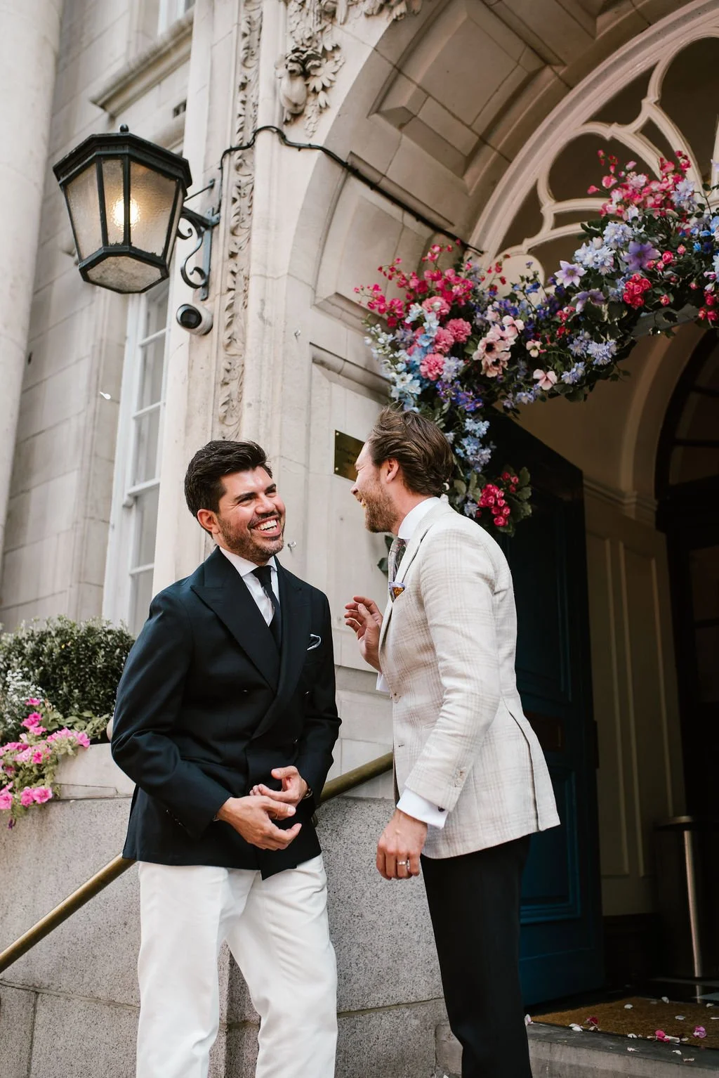 Two men smiling and talking outside a building decorated with flowers and a lantern.