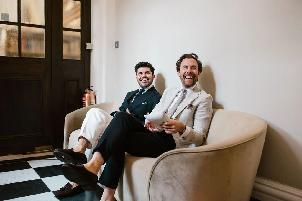 Two men sitting on a beige sofa, smiling and laughing in a room with a door and black-and-white checkered floor.