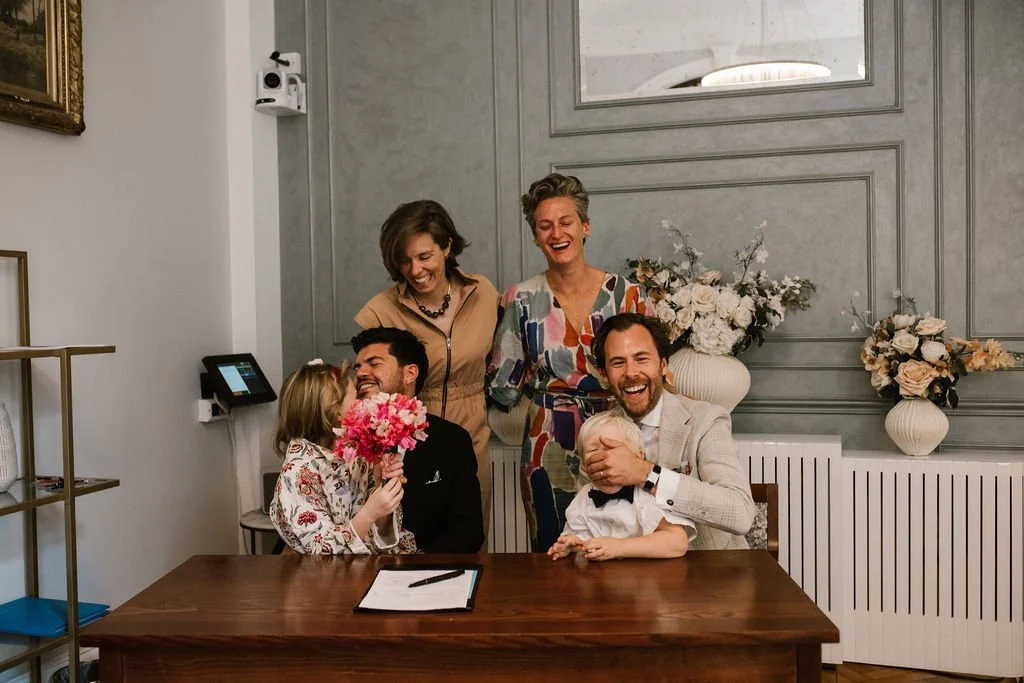 A group of six people, including two children, are gathered around a table in a warmly decorated room. They are laughing and smiling, with a woman holding a bouquet of pink flowers and a man with a beard holding a young boy. The background features large vases with white and peach flowers and a gray paneled wall.