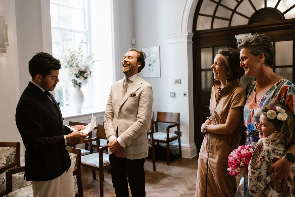 A wedding ceremony with five people, including the groom reading vows, the officiant smiling, and three women in dresses, one holding a bouquet, in a bright, elegant room with large windows and floral decorations.