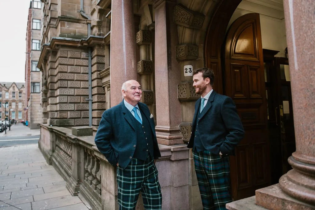 Two men in suits and tartan trousers standing outside a historic building, smiling and chatting.