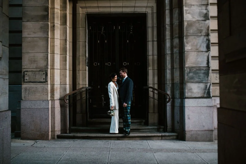 A couple stands on the steps of a building with large black gate, facing each other and smiling. The woman is holding a bouquet, and both are dressed in formal attire.