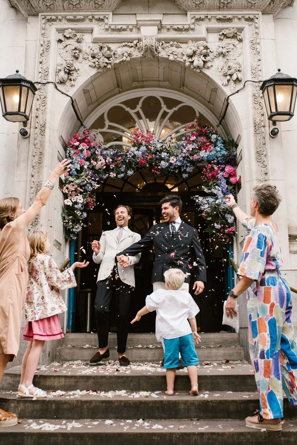 Two men in suits celebrating and holding hands on the steps of a building decorated with flowers and confetti, surrounded by cheering guests.