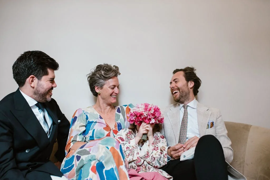 A family sitting on a beige couch, smiling and laughing, with a young girl holding a bouquet of pink flowers covering her face.