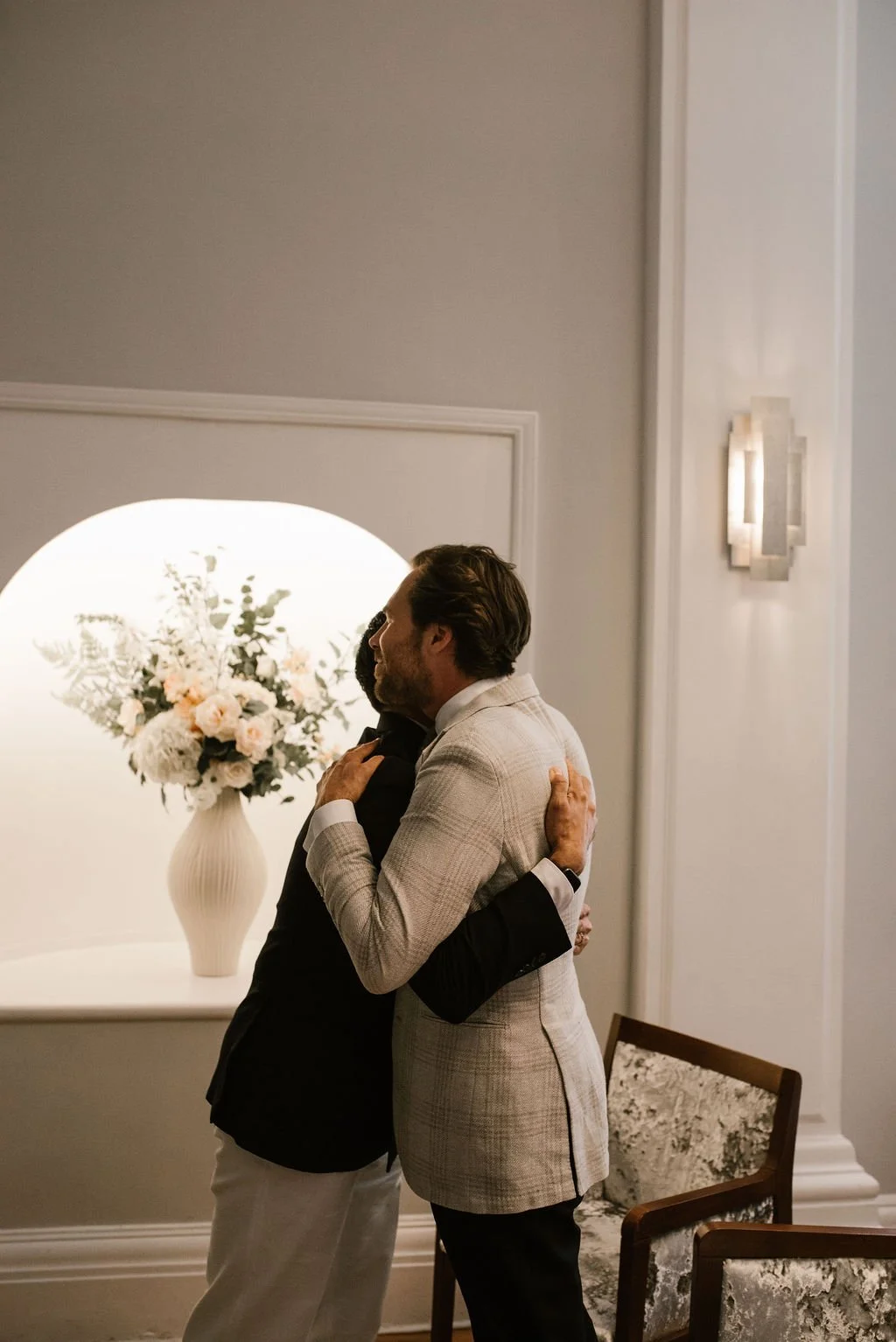Two men hugging in a warmly lit room, with a large vase of flowers on a white table and a decorative wall sconce.