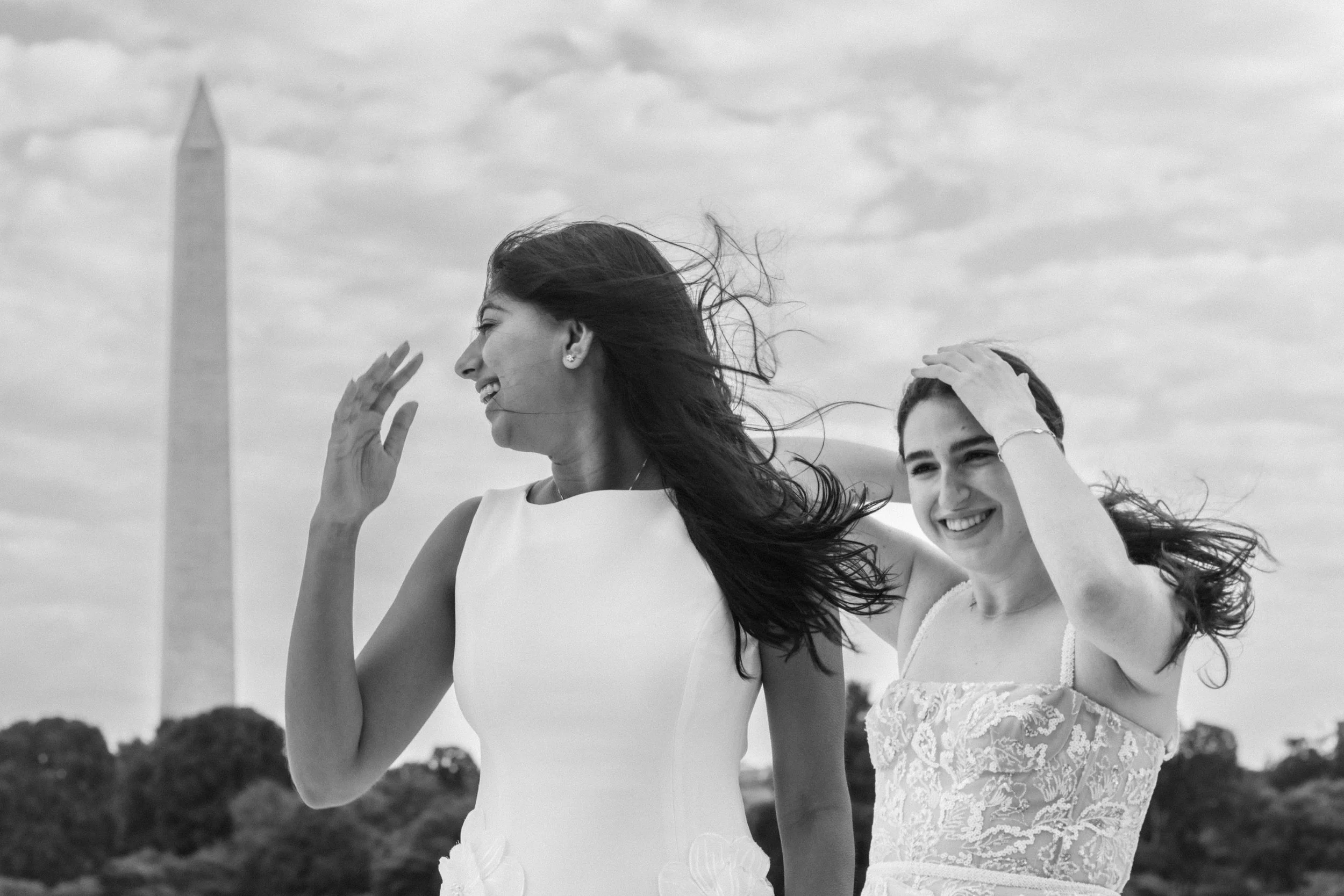 Two women smiling and laughing outdoors with windblown hair, one wearing a white dress with lace details and the other in a sleeveless dress, with the Washington Monument in the background.